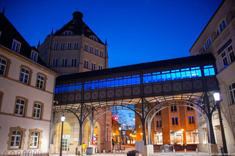 Enclosed blue-lit pedestrian bridge crosses above a street on decorative iron supports between historic buildings.