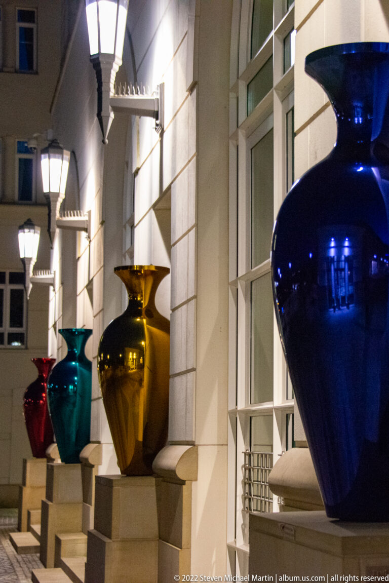 Large glossy blue, amber, turquoise, and red vases line steps beside a pale stone building at night.