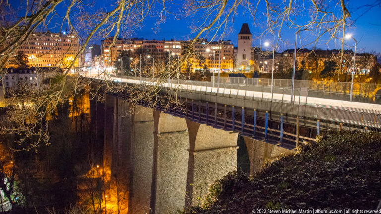 Modern bridge on tall supports crosses a deep wooded valley below illuminated upper-city buildings and a square tower.