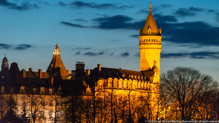 Illuminated historic complex with steep roofs, dormers, and a tall square tower capped by a pointed roof.