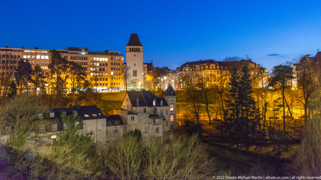 Historic lower-city houses and bare trees sit below an illuminated square tower on the upper plateau at dusk.