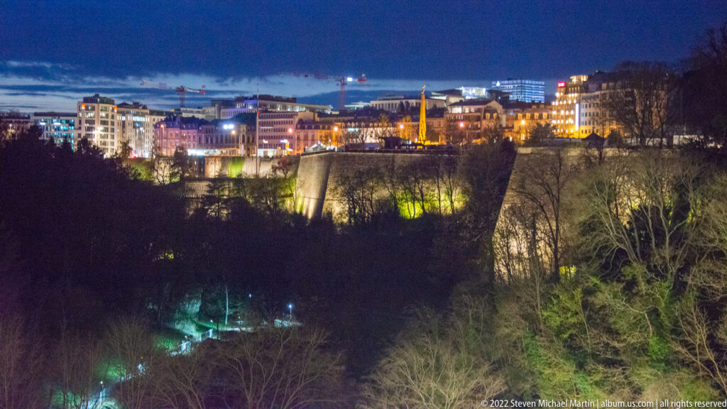 Illuminated upper-city buildings line a high fortified escarpment above a dark wooded valley at night.