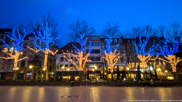 Blue- and warm-white light-wrapped trees line a paved square in front of restaurants and historic façades.