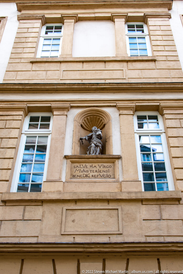Carved animal-head relief above a Latin inscription between windows on a rusticated stone façade.