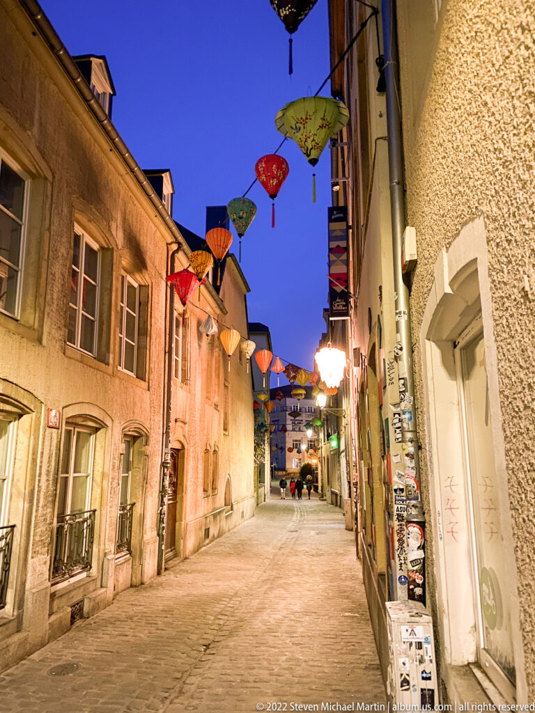 Narrow cobbled historic street with colorful parasol-like decorations hanging overhead beneath a blue evening sky.