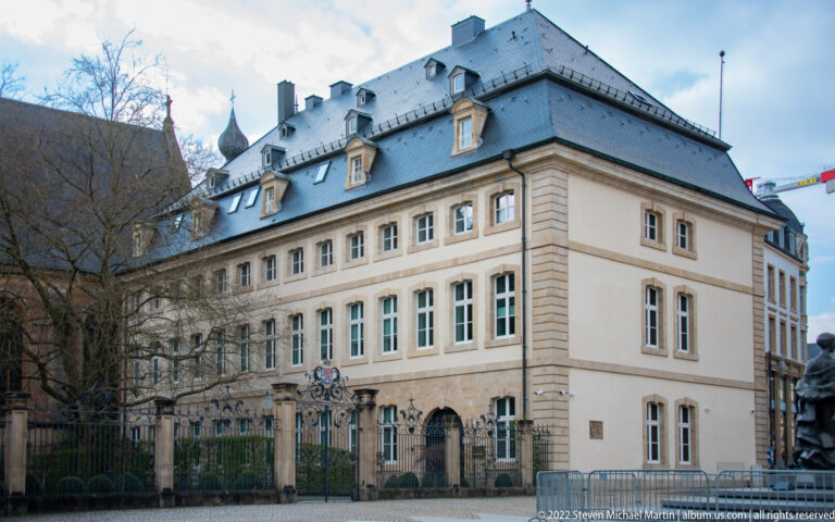 Large pale-stone government building with dormered slate roof and ornamental iron fence viewed from the street.