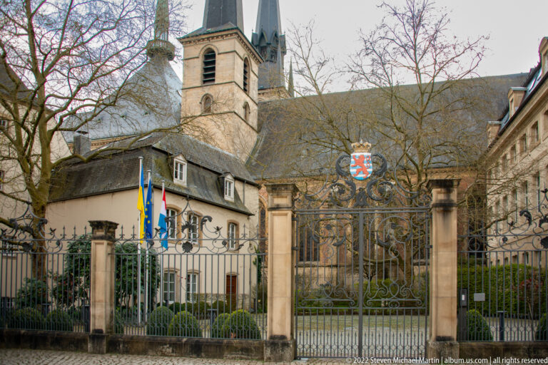 Gated historic complex with Luxembourg and European flags, heraldic crest, and a church tower rising behind.