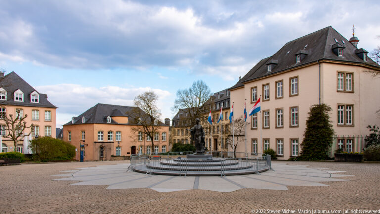 Paved civic square with circular fountain, pastel historic buildings, flags, and leafless pruned trees.
