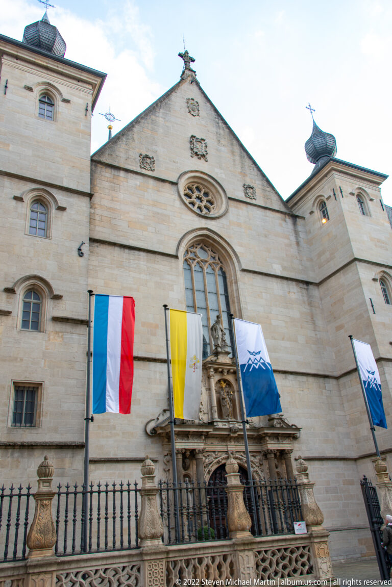 Pale stone church façade with sculpted portal, arched windows, small rose window, and vertical flags.