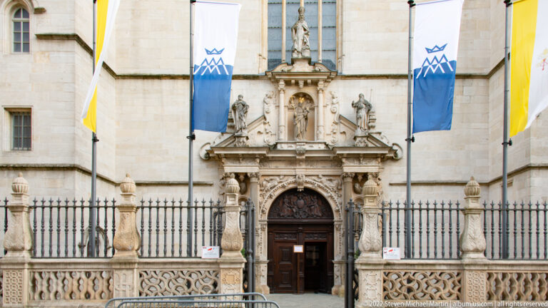 Ornate church doorway with carved saint figures, columns, balustraded stonework, and vertical banners.