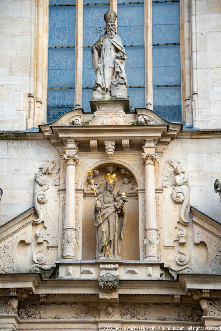 Crowned saint holding a long staff stands in an ornate niche framed by columns and carved scrollwork.