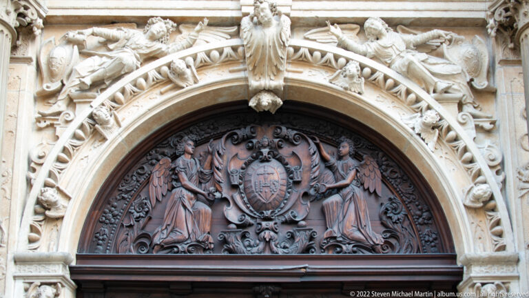 Carved wooden heraldic panel with two figures sits beneath an ornate stone arch filled with sculptural decoration.