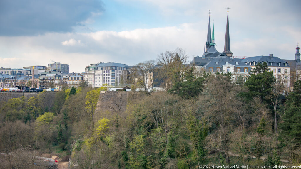 Twin church spires and upper-city buildings rise above a deep wooded ravine under a partly cloudy sky.