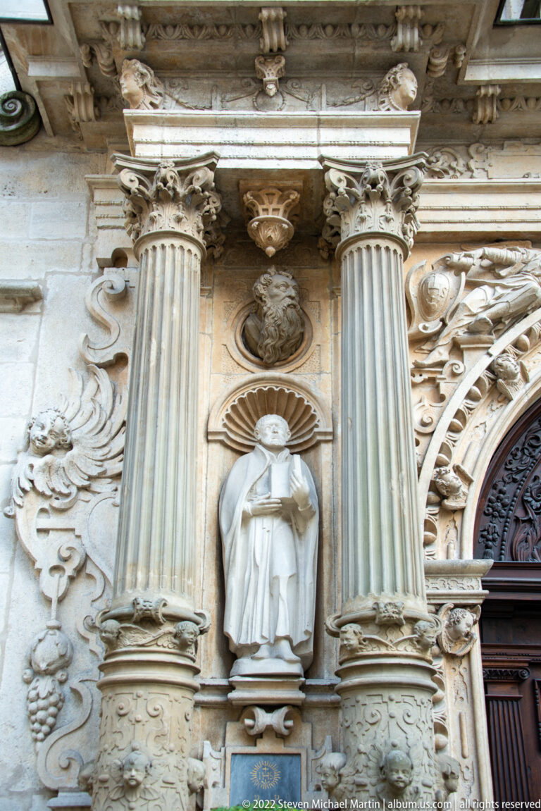 White-robed religious figure in a niche between fluted columns amid dense carved stone ornament.