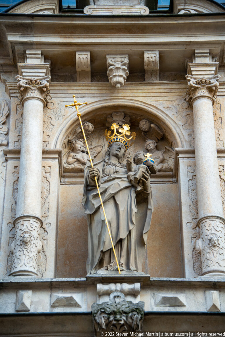 Crowned saint with a long golden staff and smaller figure stands in an ornate niche between large columns.