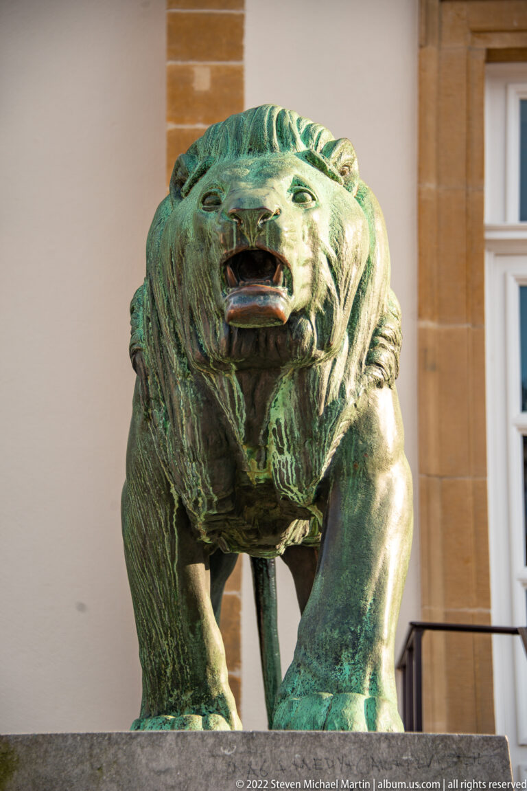 Patinated bronze lion with open mouth stands on a stone plinth outside the city hall.