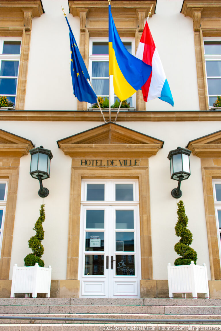 Luxembourg City Hall entrance marked 'HÔTEL DE VILLE' beneath official flags, lanterns, and topiary.