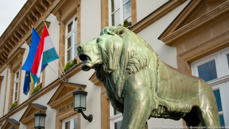 Profile of a patinated bronze lion outside city hall with Luxembourg and European flags hanging behind.