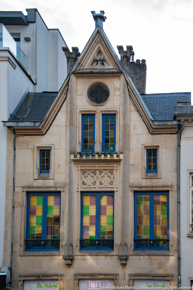 Gable-front historic building with multicolored window panels, pale stone trim, triangular pediment, and dark slate roof.