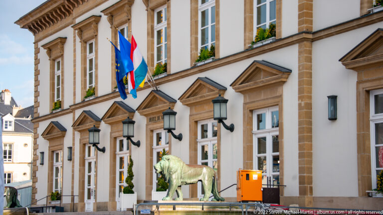Oblique view of Luxembourg City Hall with pedimented doors, flags, lanterns, and a bronze lion along the façade.