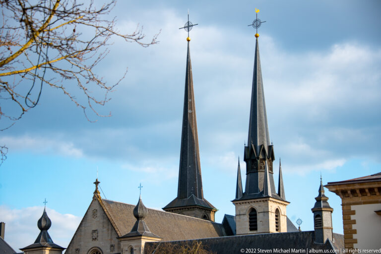 Three tall pointed church spires rise above historic rooftops beneath a blue and gray sky.
