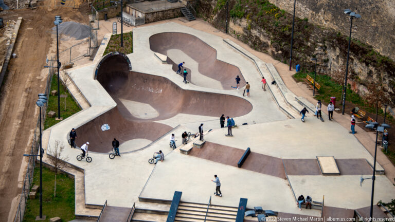 People ride and stand around a sunken concrete skatepark bordered by high historic stone walls.