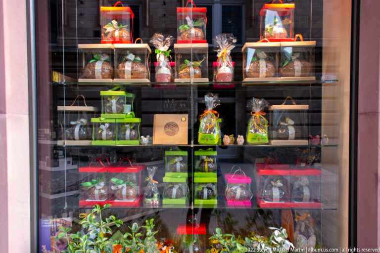 Chocolate shop window with gift-wrapped confections, boxed figures, red and green accents, and street reflections.