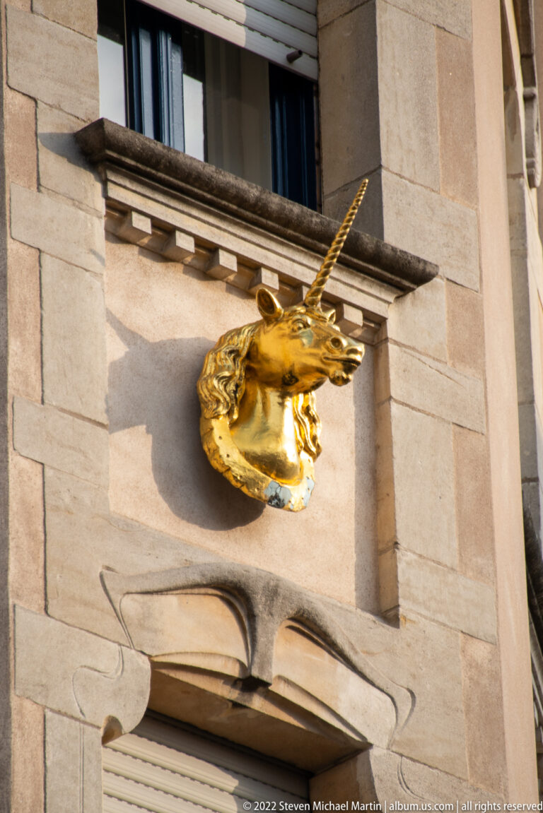Gilded unicorn head with a long spiraled horn projects from a pale stone wall above a doorway.