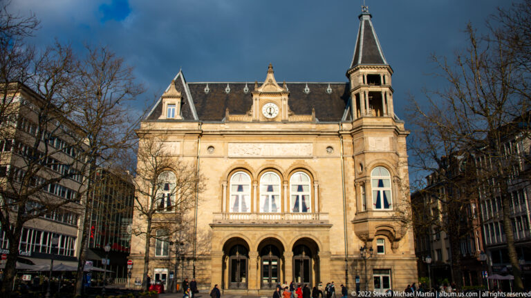 Ornate pale-stone civic building with arched entrances, central clock, steep roof, and tall corner tower.