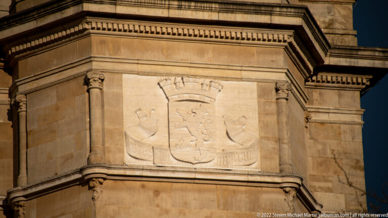 Carved heraldic shield with stylized supporters set into the corner bay of a monumental stone building.