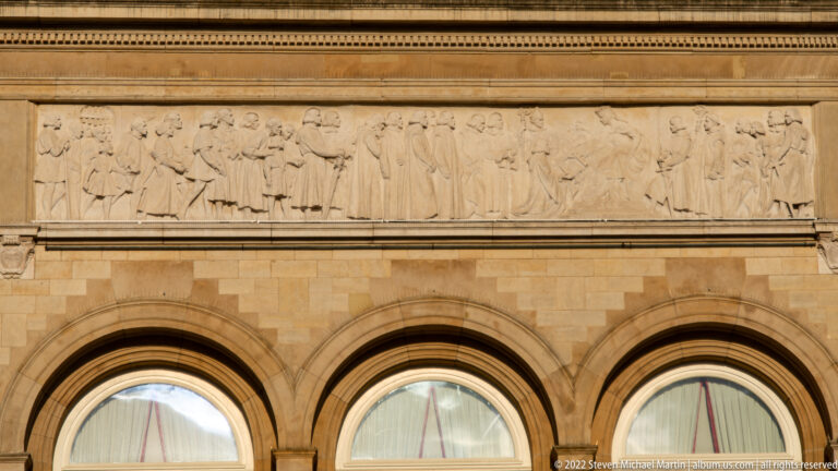 Long pale-stone figural frieze runs above three large round-arched windows on a historic public building.