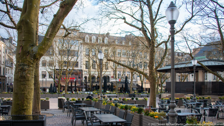 Outdoor café tables, flower beds, mature trees, and historic buildings fill a pedestrian city square.