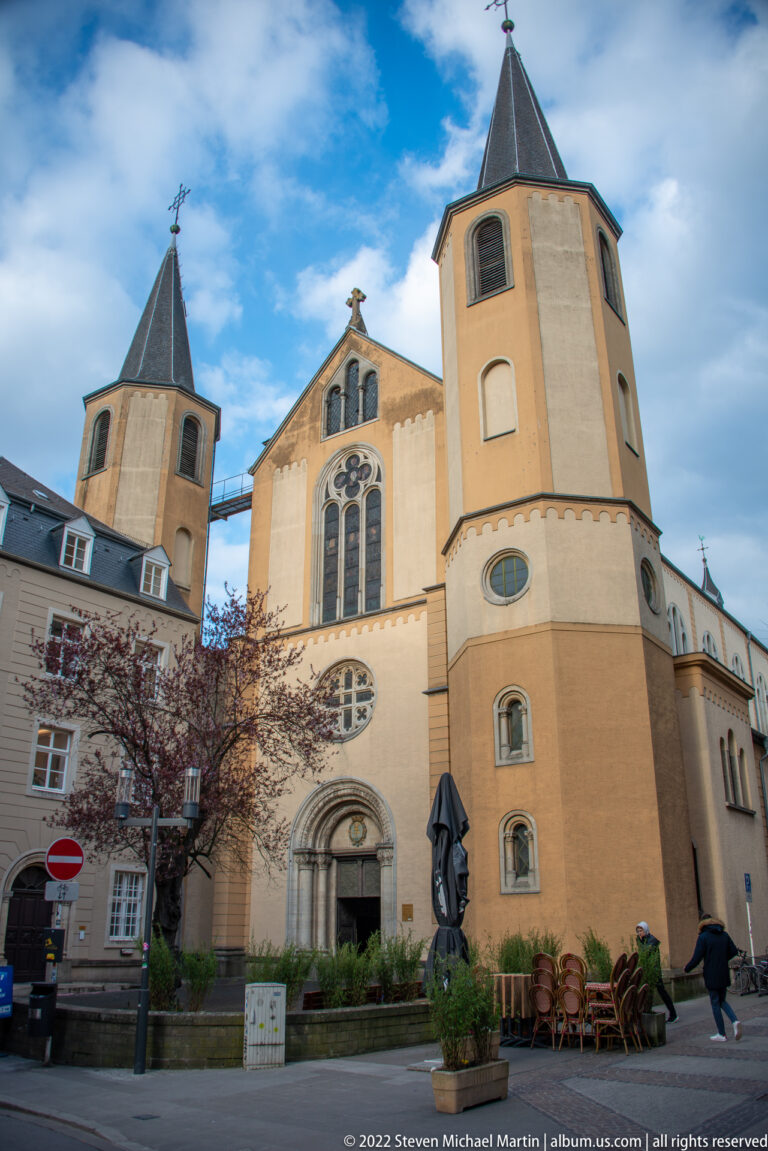 Historic pale church with twin polygonal corner towers, dark roofs, traceried windows, and a central sculpted portal.