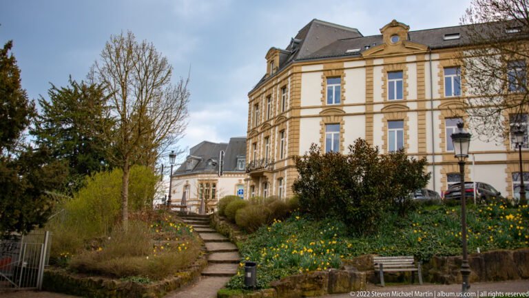 Historic cream-colored institutional building beside a stepped garden path lined with spring flowers and shrubs.