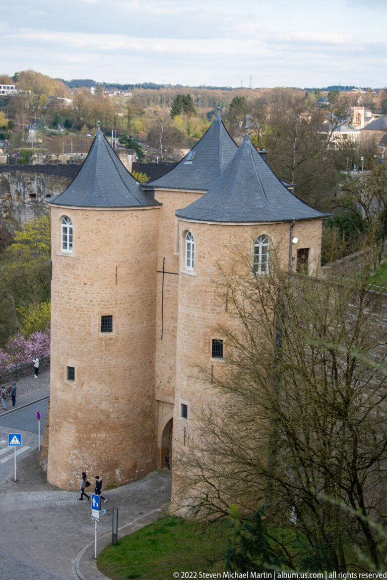 Two adjoining round masonry towers with dark conical roofs and small openings stand beside a paved urban area.