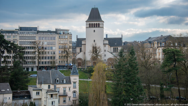White square church tower with a dark pyramidal roof rises among villas, trees, and modern apartment blocks.
