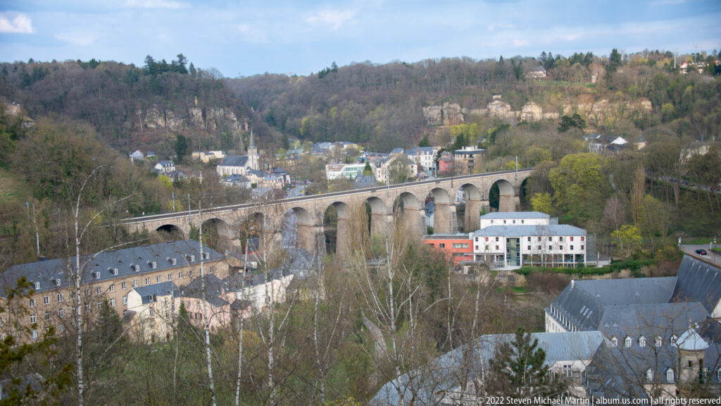 Long multi-arched stone viaduct crosses a wooded urban valley above clustered buildings and slate roofs.