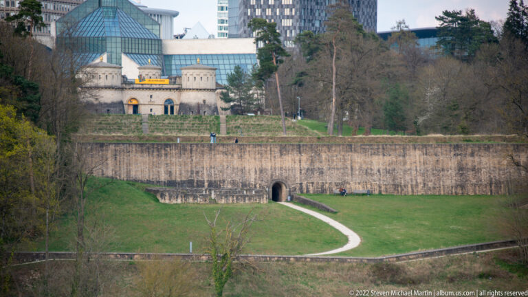 Massive stone defensive wall with an arched opening crosses a grassy slope beneath higher fortified structures.