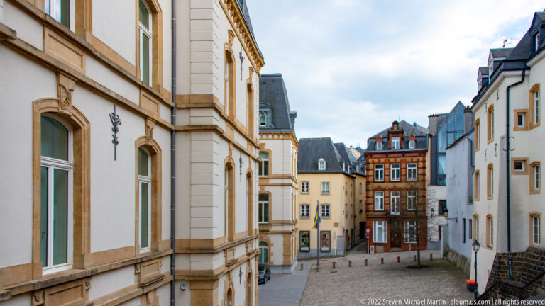 Narrow cobbled street lined with pale historic façades descends toward a dark timber-fronted building.