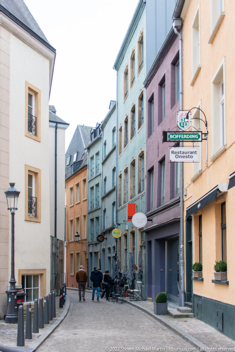 Narrow old-city street lined with pastel townhouses, restaurant signs, café furniture, and slate-roofed upper stories.
