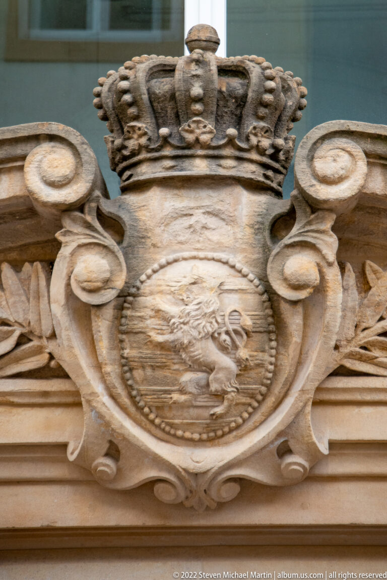 Carved crowned coat of arms in an ornate stone cartouche with scrolls and foliage above a historic façade.