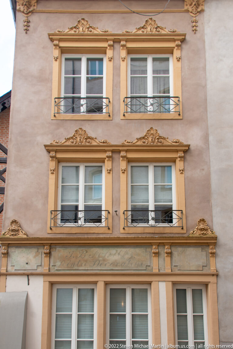 Historic townhouse façade with paired windows, decorative stucco surrounds, floral motifs, and small iron railings.