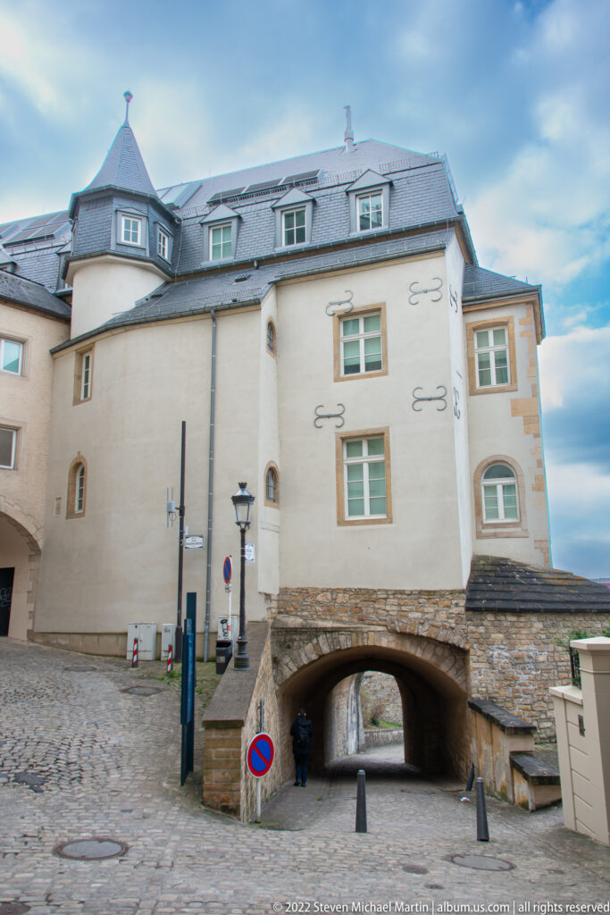 Historic pale building spans a narrow arched passage above cobbles and stone retaining walls.