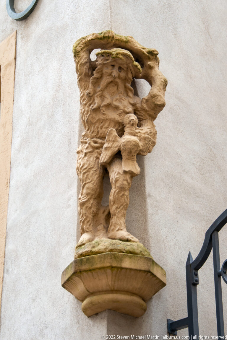 Weathered bearded stone figure projects from a wall with raised arms supporting a flat architectural element.