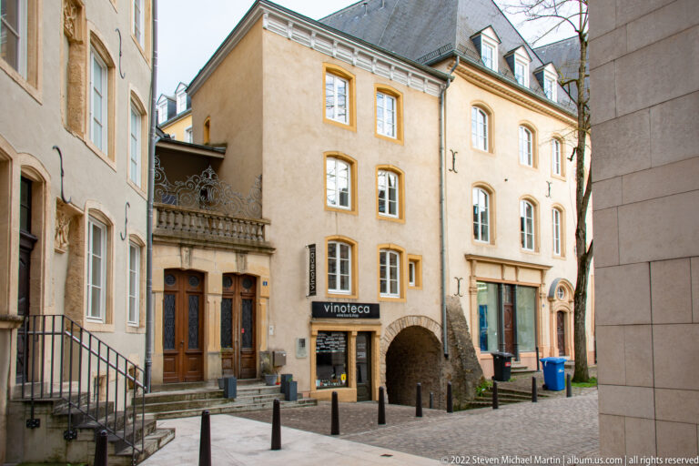 Small paved courtyard lined by historic townhouses, an iron balcony, a vinoteca storefront, and an arched passage.