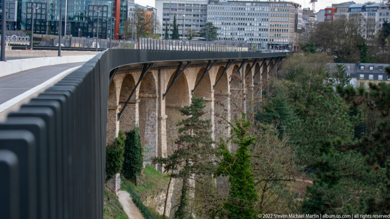 Series of tall stone viaduct arches spans a wooded ravine beneath a roadway with a dark modern parapet.