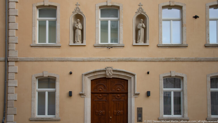 Central wooden doorway beneath two upper-story saint statues in niches with cross-shaped finials.
