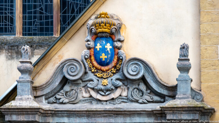 Blue shield with gold fleur-de-lis beneath a gilded crown set in an ornate stone cartouche.