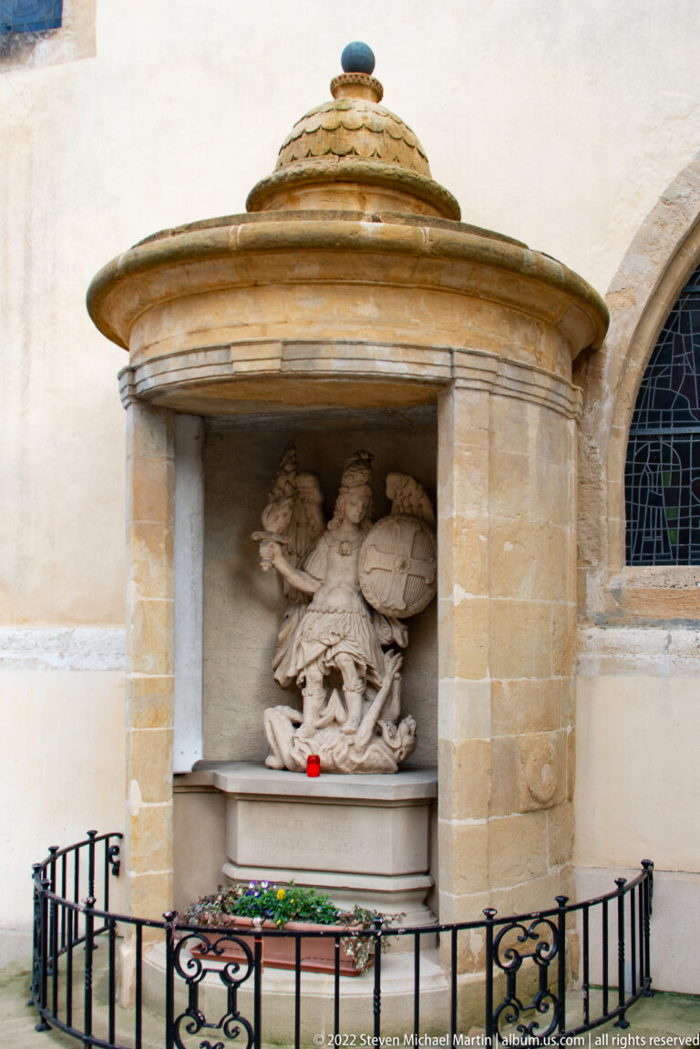 Exterior church shrine under a domed canopy contains a sculpture of Saint Michael overcoming a fallen figure.