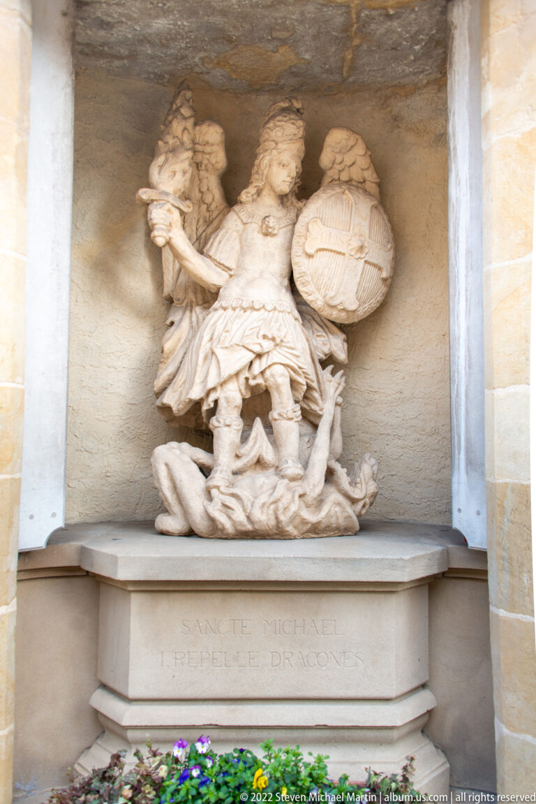 Stone sculpture of Saint Michael defeating a dragon above a pedestal inscribed with 'SANCTE MICHAEL.'
